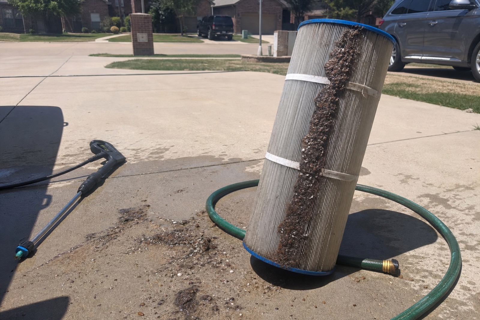 A cylindrical pool cartridge filter being rinsed in a driveway with a pressure washer, debris washed off onto concrete.